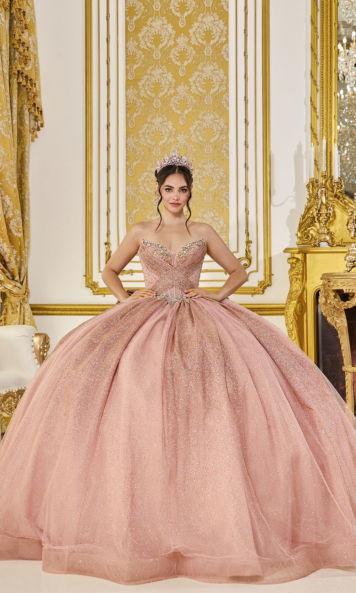 A young woman wears the Quinceanera Dress 5078 By Dancing Queen in rose gold, featuring a strapless corset bodice and tiara, as she stands in an ornate gold and white room with a gilded chair and mirror.